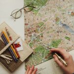 A hand writes a travel itinerary over a map with glasses and coloured pencils in a notebook