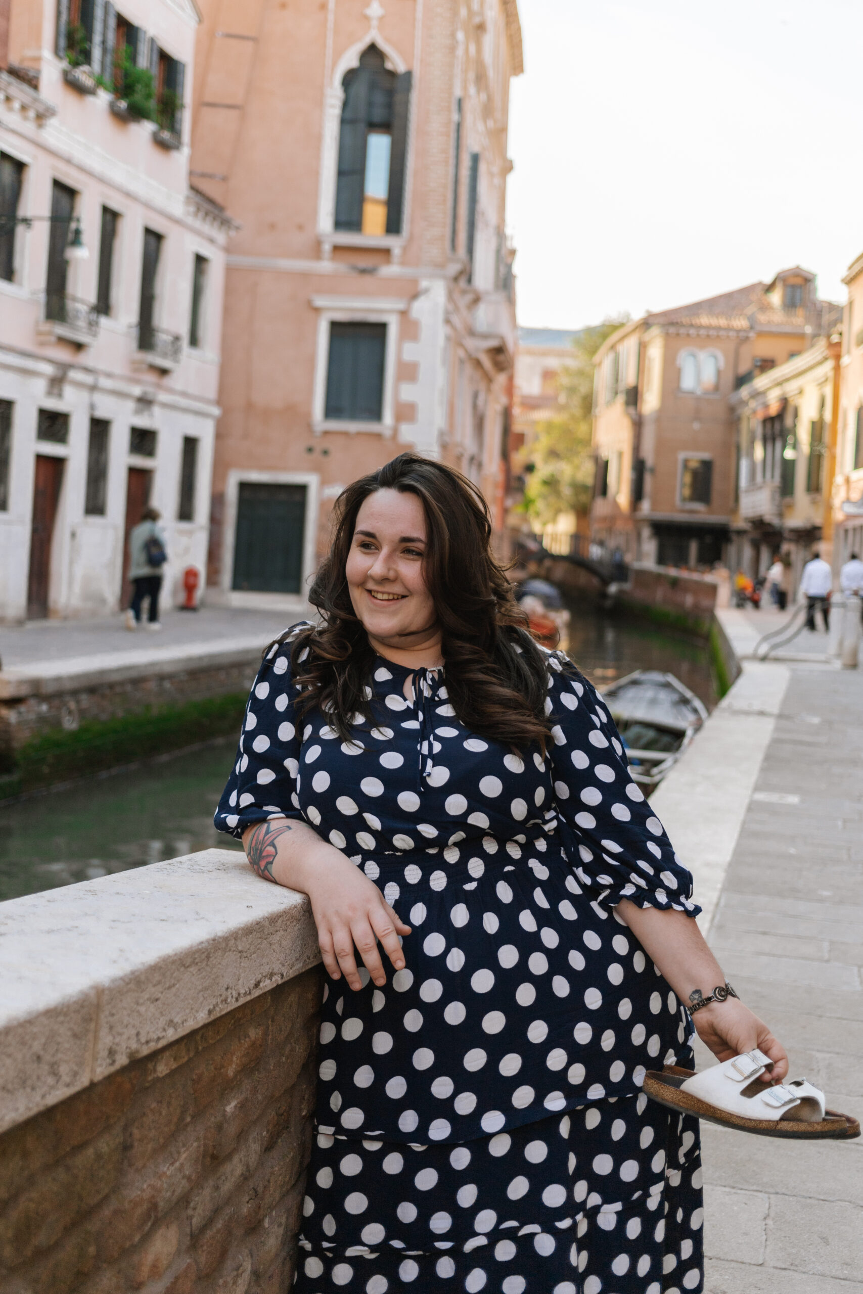 Alex wears a spotty dress and stands barefoot by a Venetian canal, smiling
