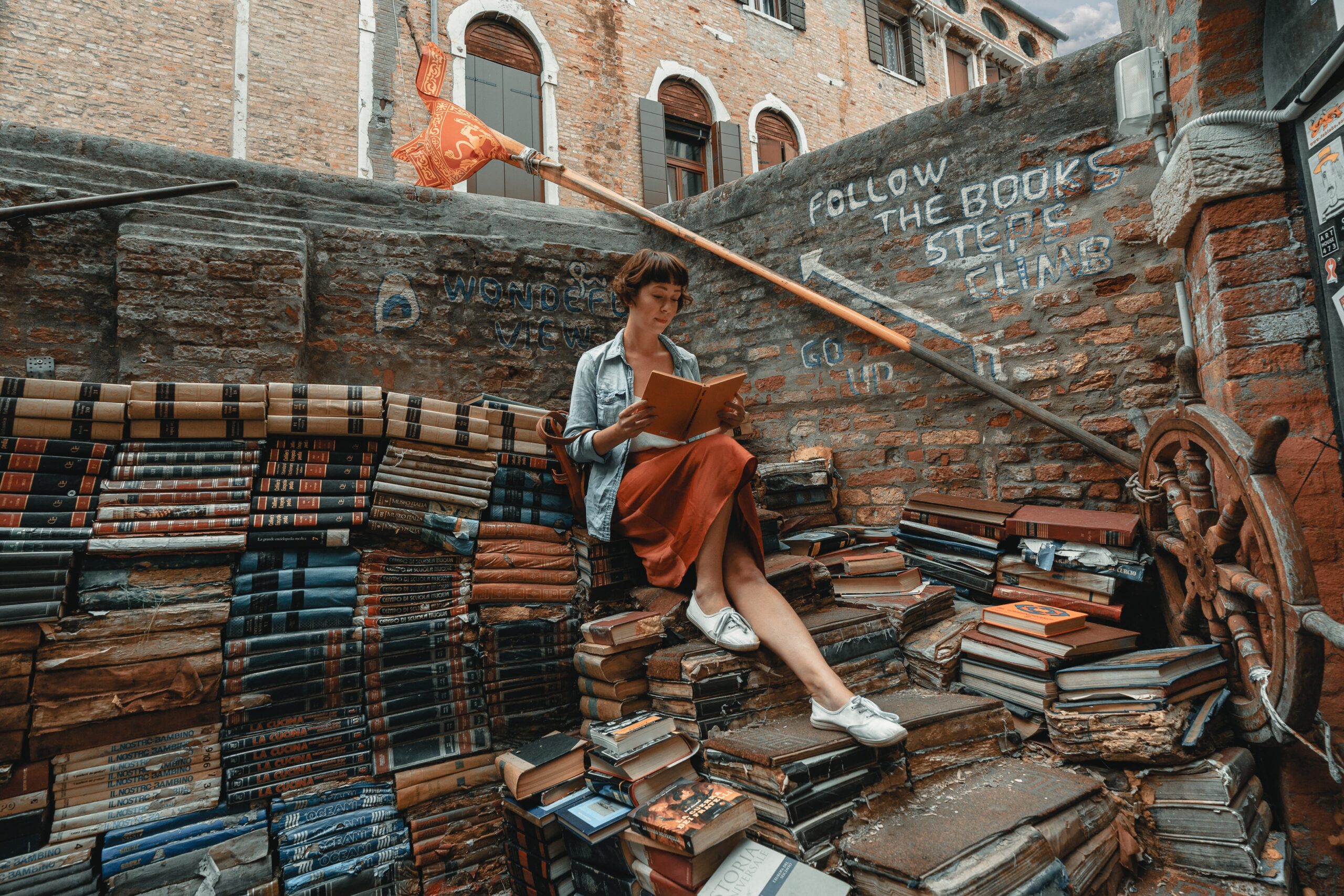 A woman sits reading a book on a staircase made from old books.